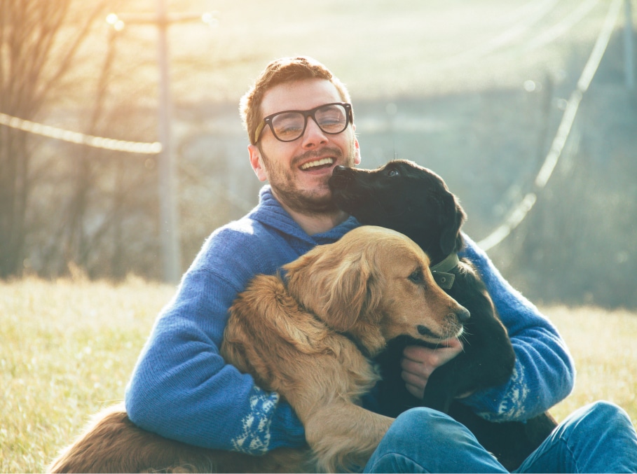 A man wearing glasses and a blue sweater sits outdoors on grass, smiling while holding a black dog and a golden retriever.