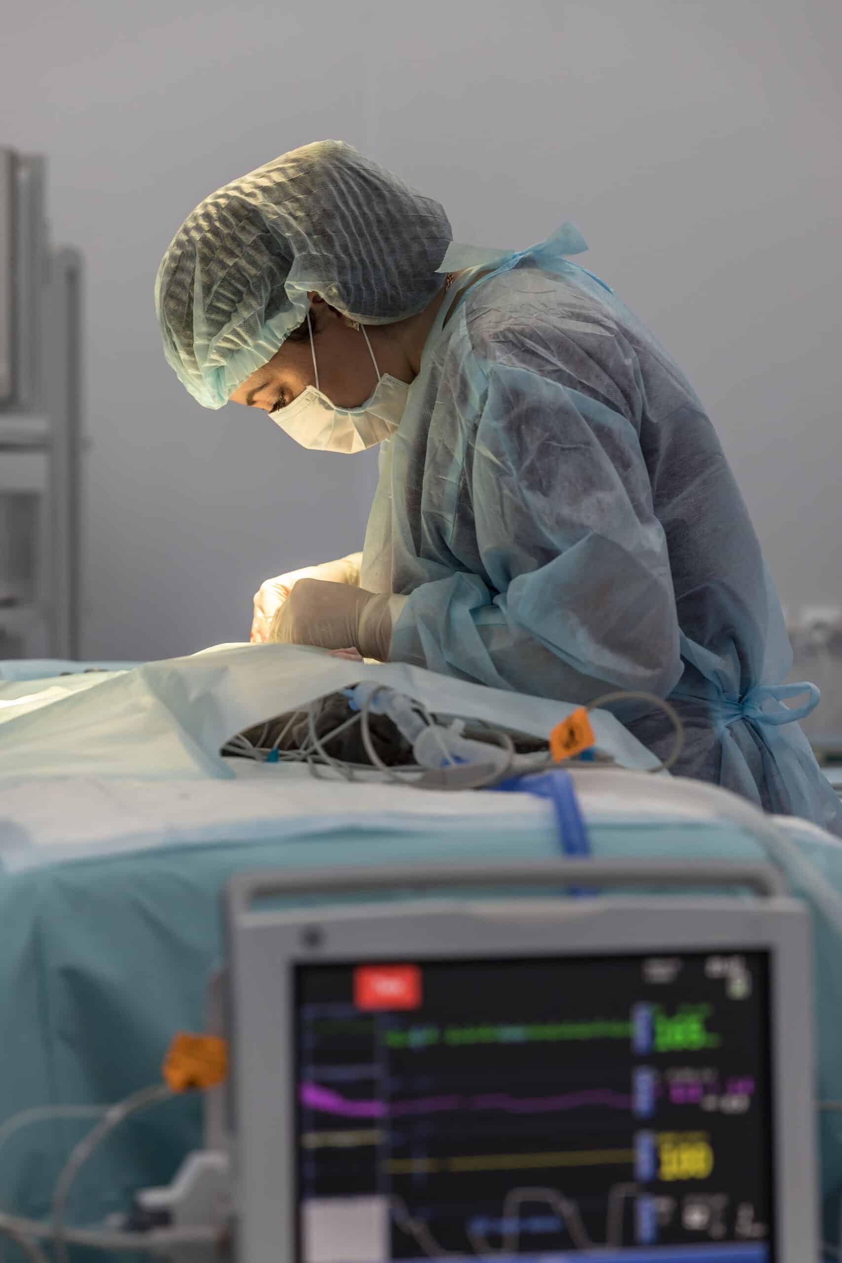 A medical professional in surgical attire performs an operation, with monitoring equipment displaying vital signs in the foreground.